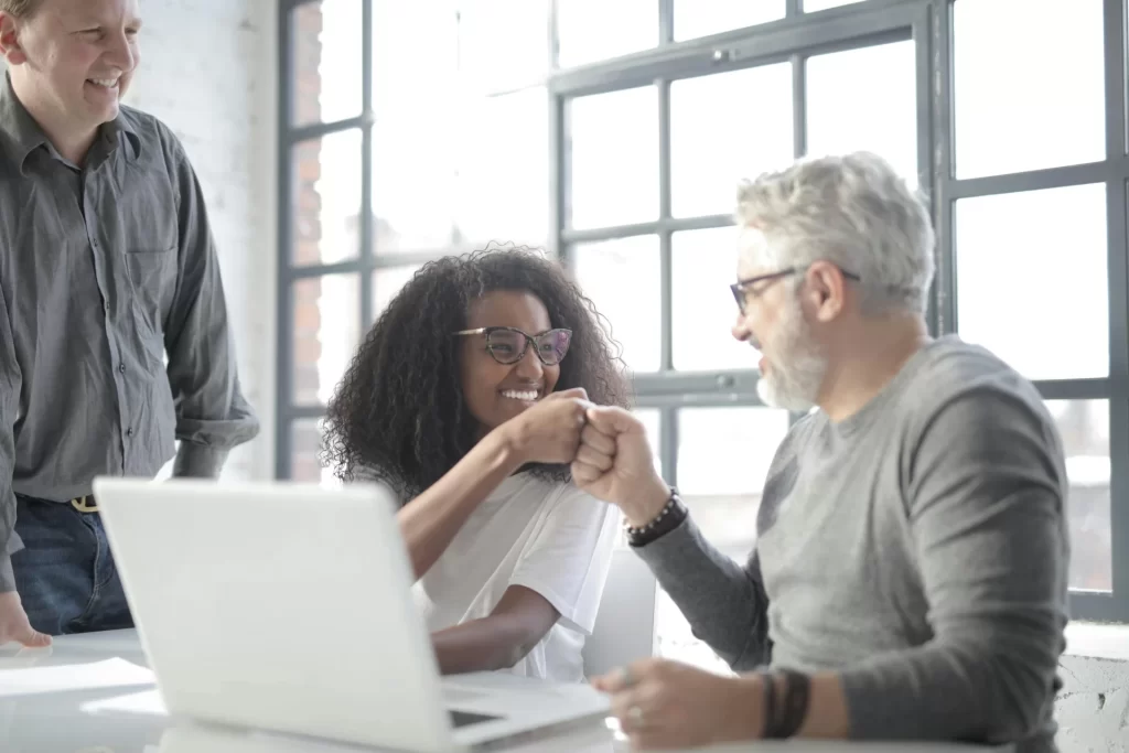 Two cheerful coworkers celebrate progress with a fist bump while reviewing work together at a laptop in an office. Another coworker stands beside them, smiling.